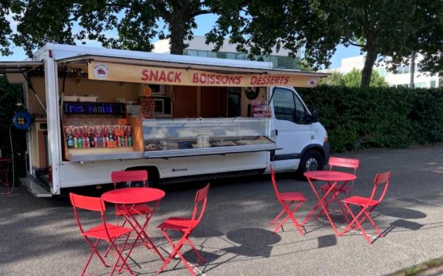 Le camion Buena Aventura installé sur un emplacement, auvent « Snack Boissons Desserts » ouvert, tables et chaises rouges dressées devant.