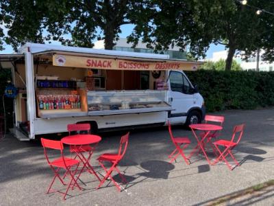 Le camion Buena Aventura installé, auvent « Snack Boissons Desserts » ouvert, tables et chaises rouges dressées devant.