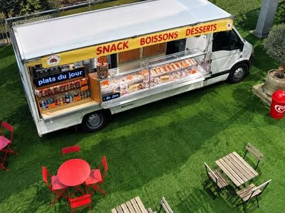 Vue du dessus du camion ouvert sur une pelouse : comptoir, vitrine, écran « plats du jour » et tables rouges et en bois dressées autour.